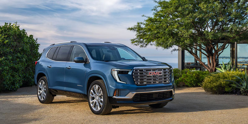 Blue GMC SUV with a bold front grille, parked on a gravel surface with greenery in the background.